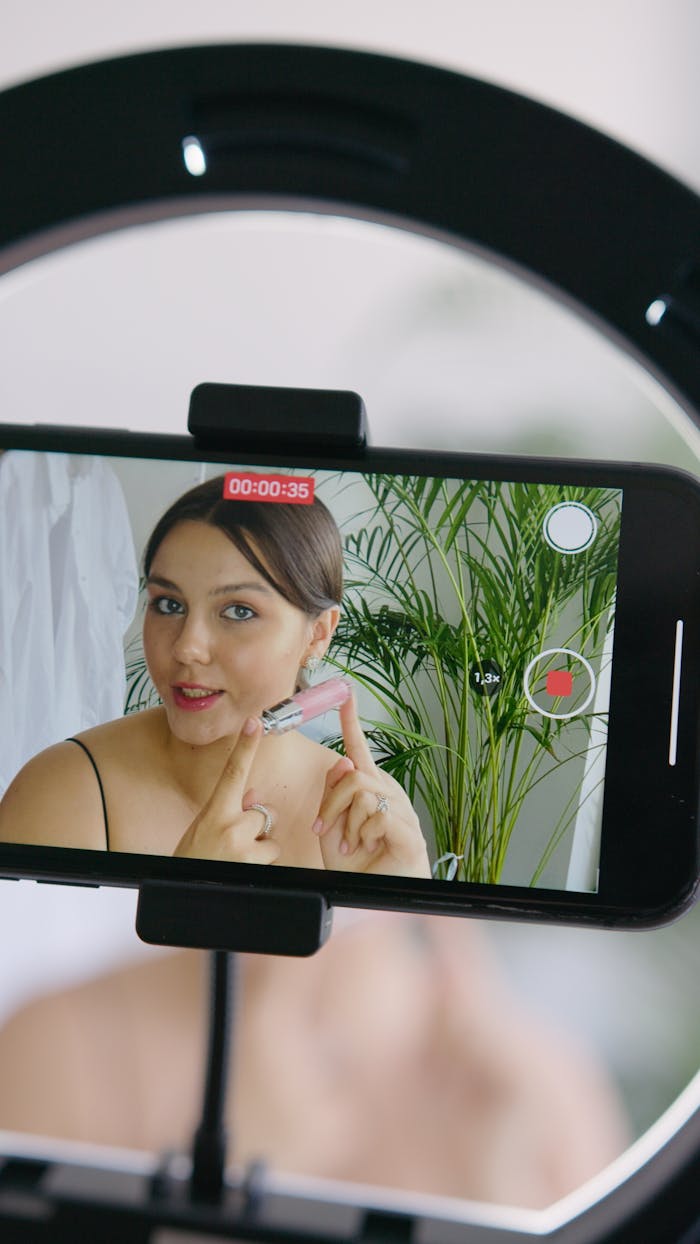services-03 A young woman records a makeup tutorial on her smartphone at home, showcasing a lip product.