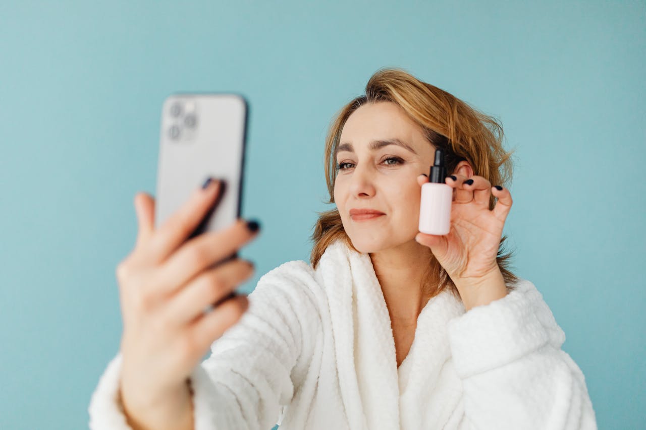 Home Woman in bathrobe taking a selfie with a cosmetic bottle against a blue background.