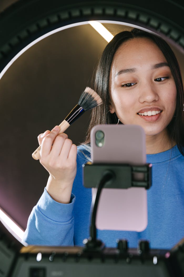services-02 Young woman streaming a beauty tutorial using a ring light and mobile phone, applying makeup indoors.