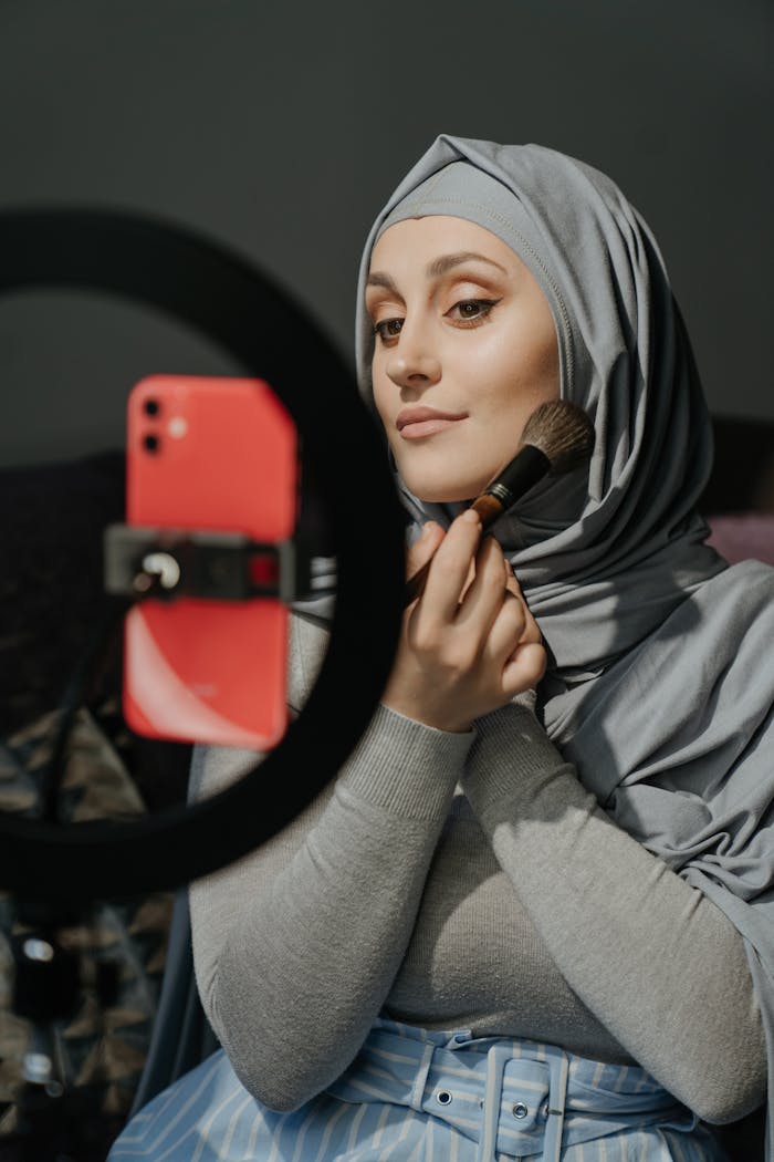 Home A woman in a hijab applies makeup while filming a beauty tutorial indoors.
